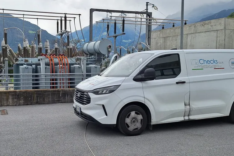Furgone CHECKS S.p.A. parcheggiato davanti a una sottostazione elettrica, con le montagne sullo sfondo